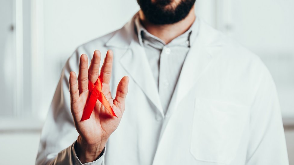 Imagen de un médico vestido de blanco, sosteniendo una cinta roja de concienciación de VIH