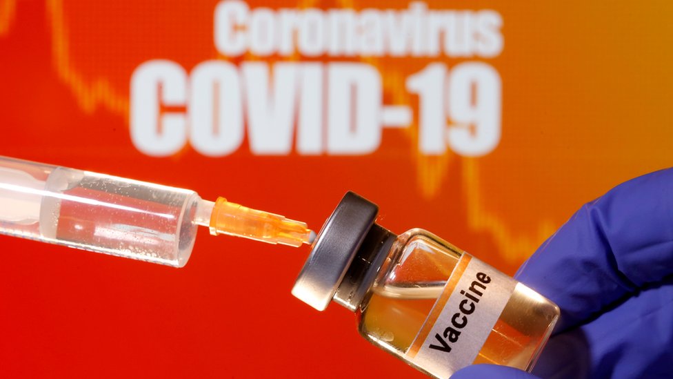 A small bottle labeled with a "Vaccine" sticker is held near a medical syringe in front of displayed "Coronavirus COVID-19" words in this illustration taken April 10, 2020