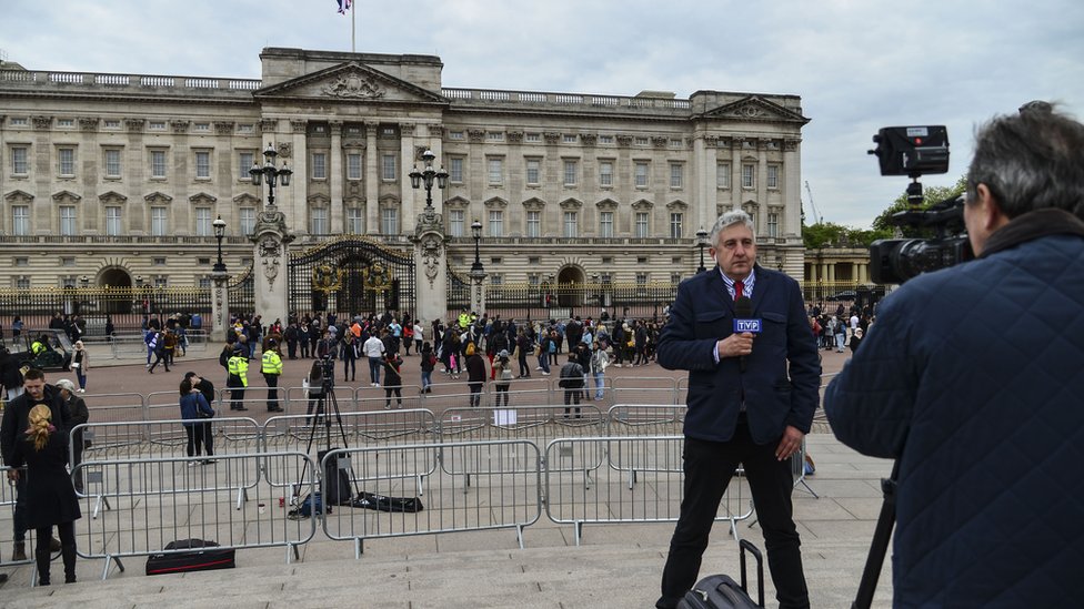 A news broadcaster speaks to camera as members of the public queue to read the official notice of the birth of a baby boy to the Duke and Duchess of Sussex outside Buckingham Palace on May 6, 2019