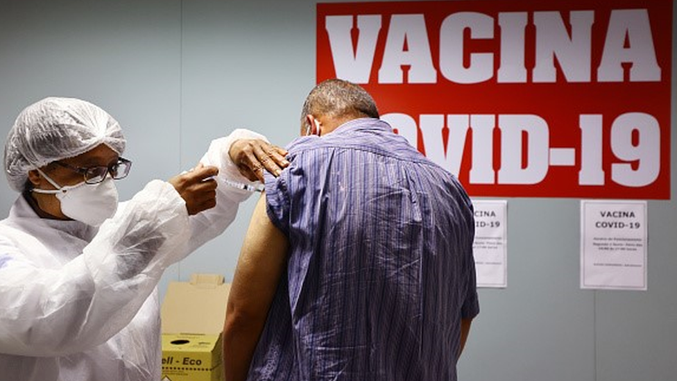 Enfermeira aplica vacina no braço de homem numa sala com um cartaz escrito 