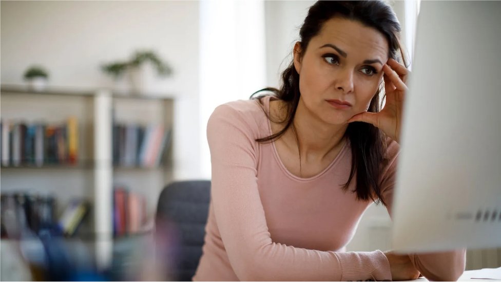 Mulher branca de cabelo preto está sentada em frente ao computador com a mão nas têmporas