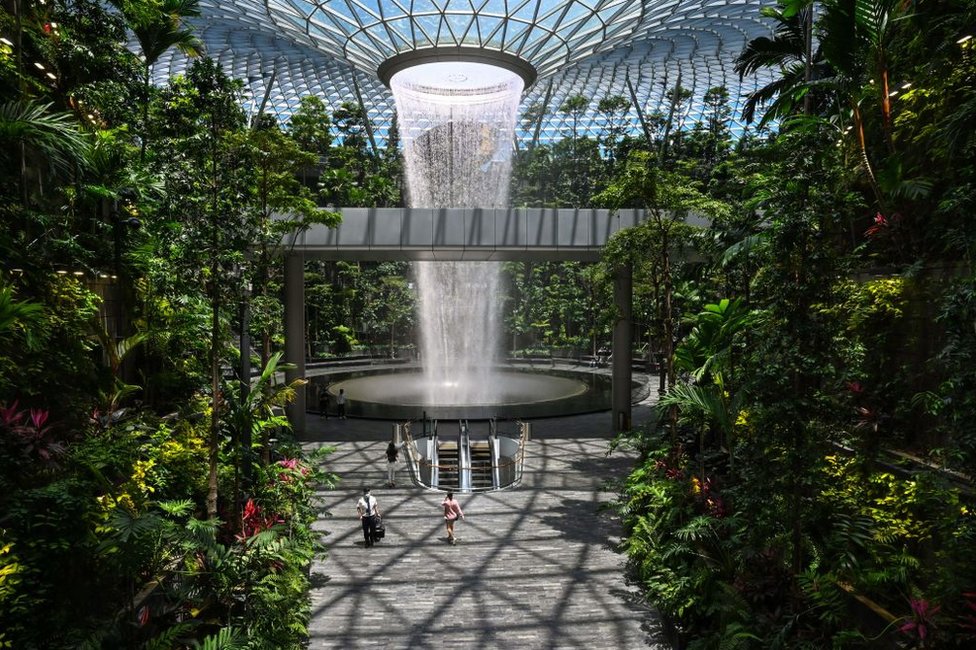 ما الذي يجعل مطار تشانغي السنغافوري الأفضل على مستوى العالم؟ 2 People walk past the Rain Vortex indoor waterfall feature at Jewel Changi Airport in Singapore on August 19, 2021.