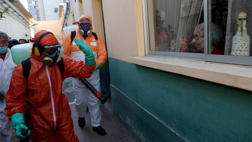 Рабочий в защитном снаряжении машет женщине, используя дезинфицирующее средство для уборки за пределами дома в Сантьяго, Чили