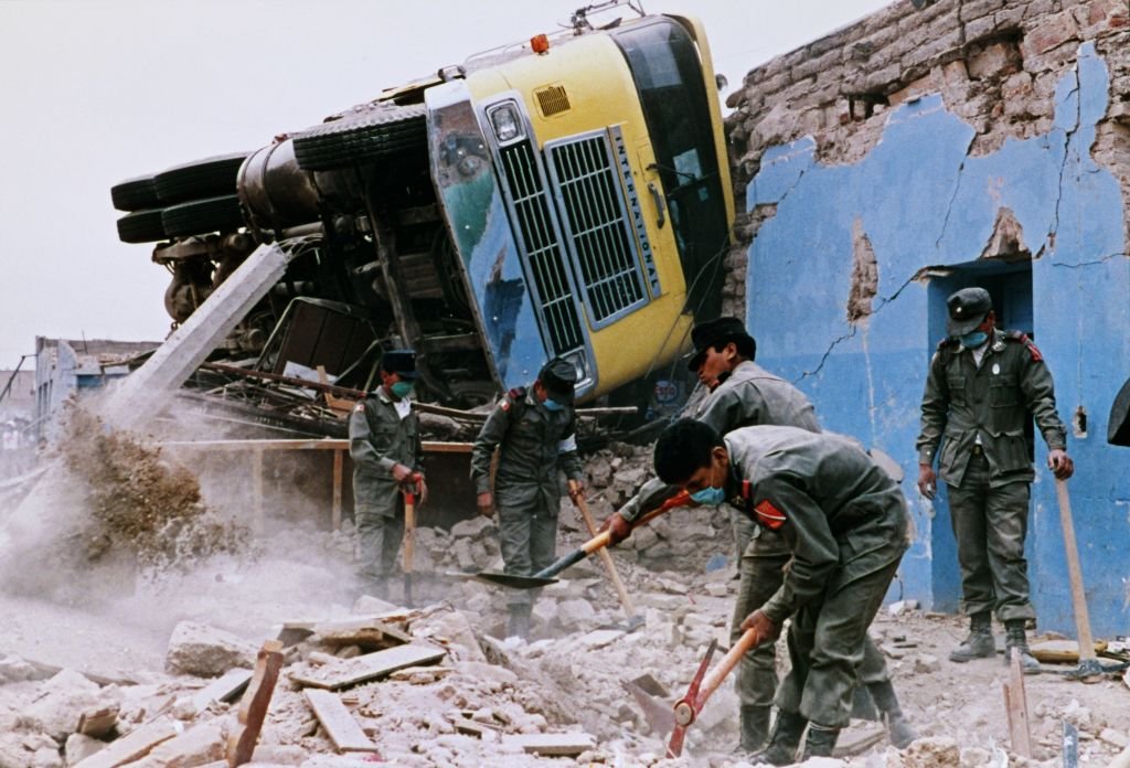 Un bus volteado y casa destruida con dos hombres ayudando