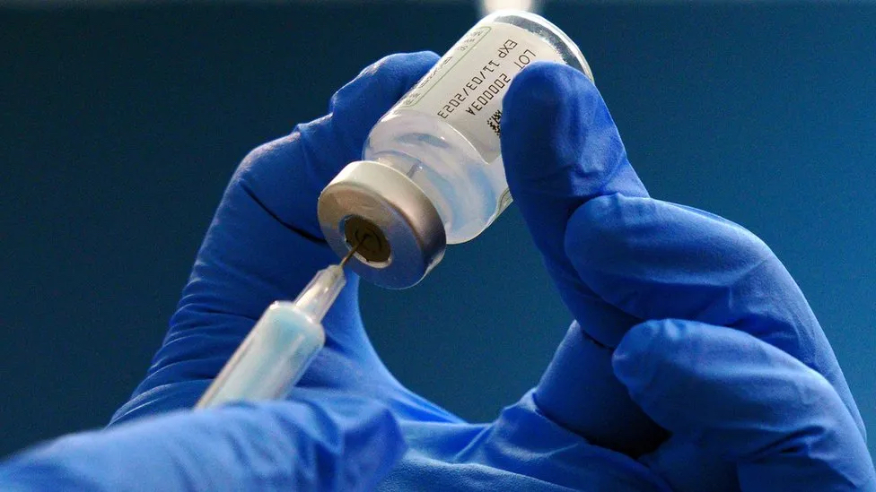 Two hands in gloves holding up a syringe needle going into a vaccine vial top