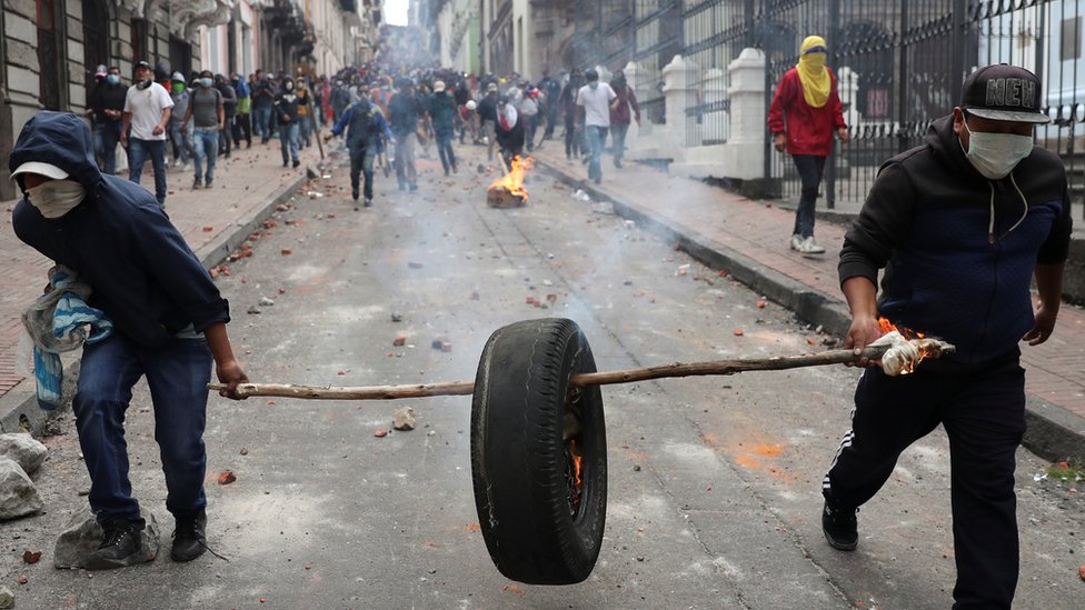 Протестующие несут горящую шину по дороге, усыпанной камнями