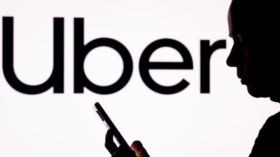 A woman in silhouette holds a smartphone with the Uber logo in the background.