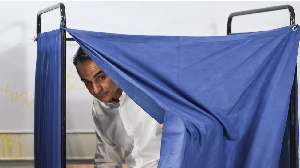 Кириакос Мицотакис выглядывает из-за синей занавески кабины для голосования