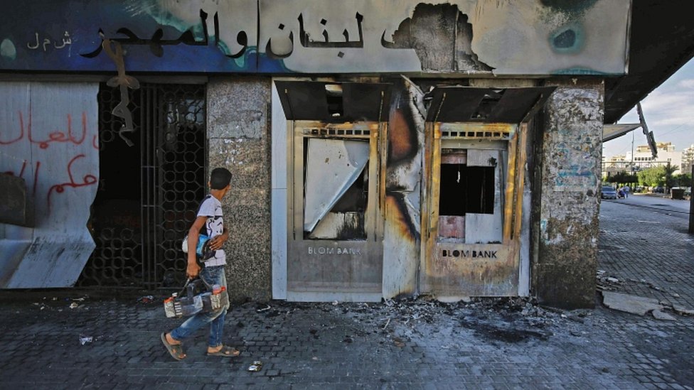 Молодежь проходит с комплектом для чистки обуви мимо сгоревшего отделения ливанского банка после того, как оно было подожжено и разгромлено протестующими ранее, на площади Аль-Нур в северном портовом городе Ливана, Триполи, 12 июня 2020 года