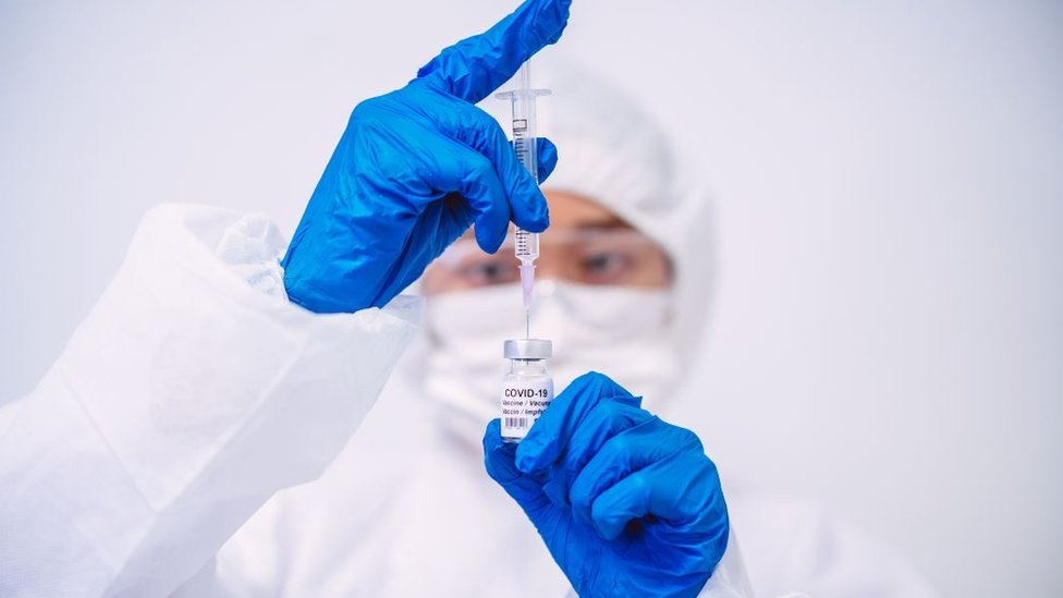 Доктор в защитных перчатках и спецодежде заполняет шприц для инъекций вакциной Covid-19 - стоковое фото
