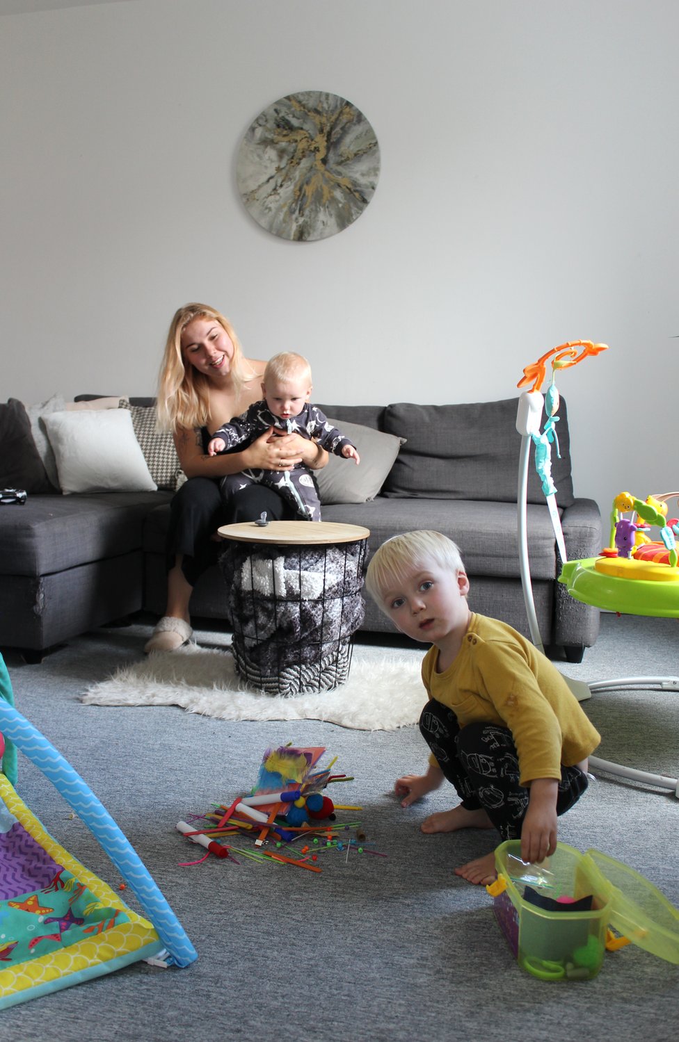Мать сидит на диване, держа своего ребенка, пока ее малыш играет со своими игрушками на полу