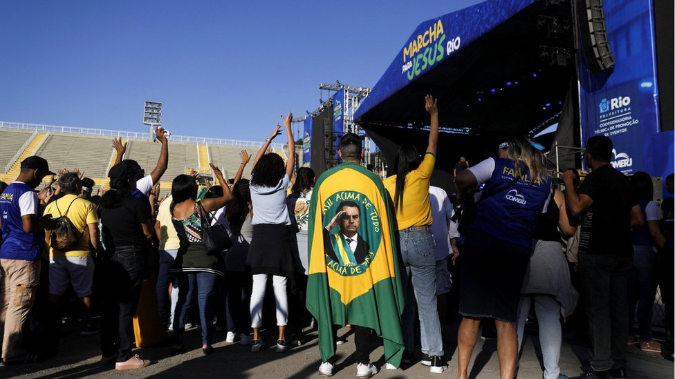 Fotografia colorida mostra um homem de costas enrolado em uma bandeira do Brasil em que no centro há uma foto de Bolsonaro; o homem está ao lado de algumas pessoas em frente a um palco onde está escrito 