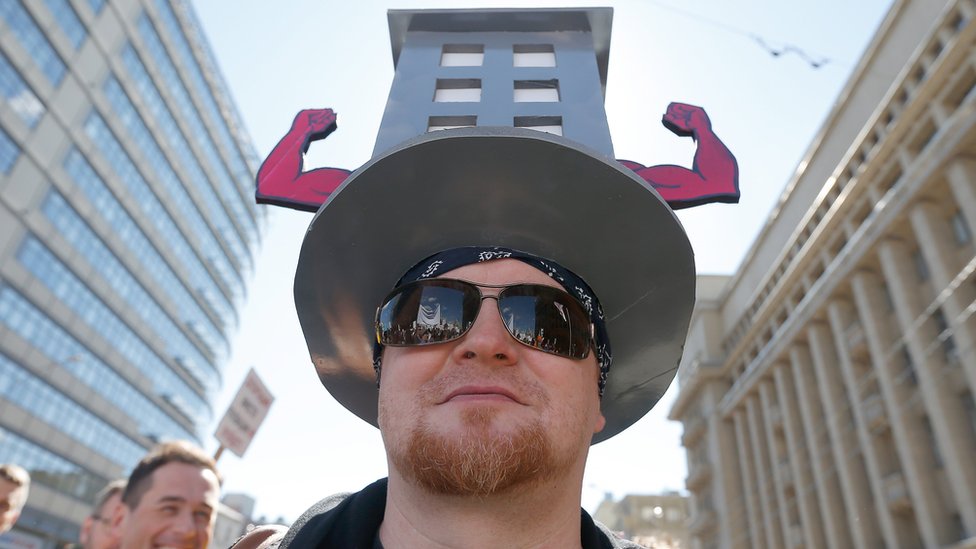 Протестующий в шляпе с картонным вырезом невысокого дома, с торчащими по бокам мускулистыми согнутыми руками