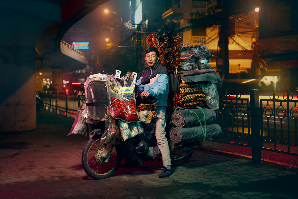 Мужчина изображен на мотоцикле, который завален автомобильными аксессуарами, такими как коврики и вышитый бисером чехол на сиденье