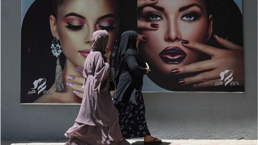 Dos mujeres caminan frente a una valla que promueve un salón de belleza en Kabul, agosto 2021