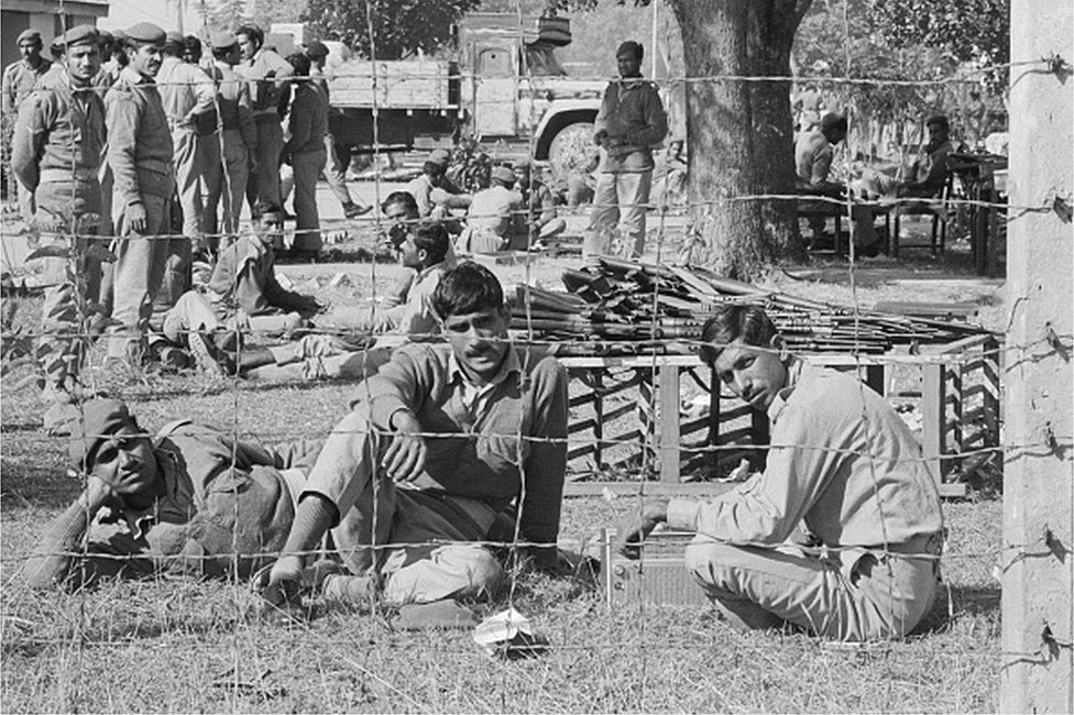 Пленные пакистанские солдаты сидят за забором из колючей проволоки в лагере для военнопленных.