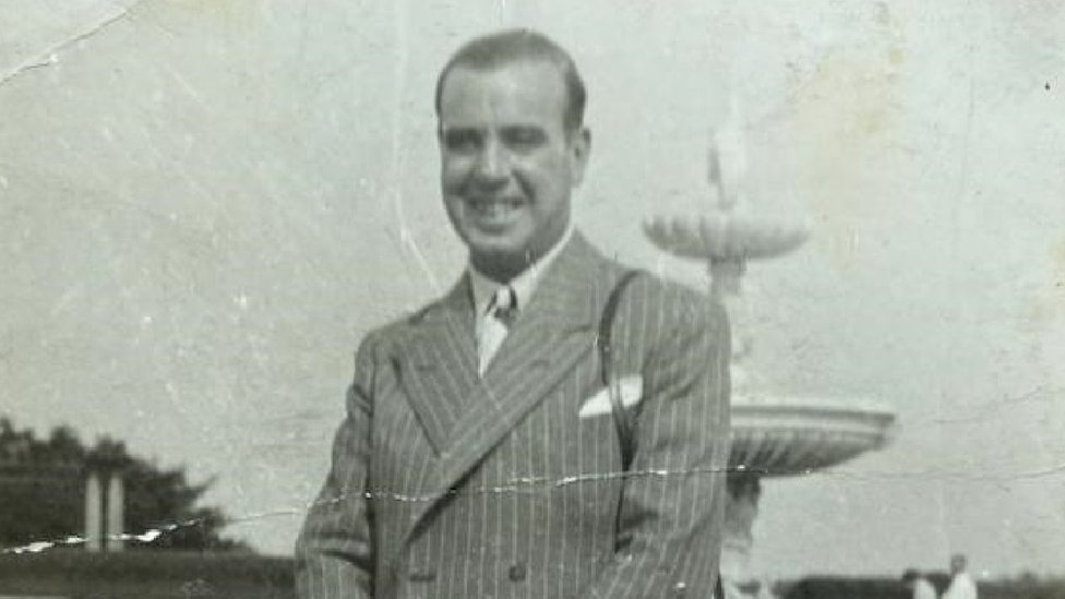 Una foto antigua en blanco y negro de un hombre con traje y una cámara colgada del hombro