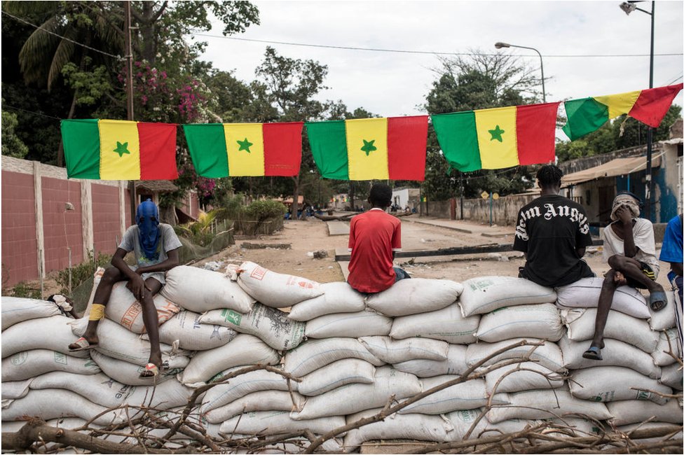 متظاهرون يجلسون على حواجز من أكياس الرمل تحت الأعلام السنغالية.
