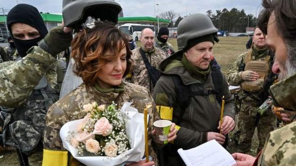 Soldado segura o capacete de Lesya acima da cabeça para proteger seu véu de casamento durante a cerimônia