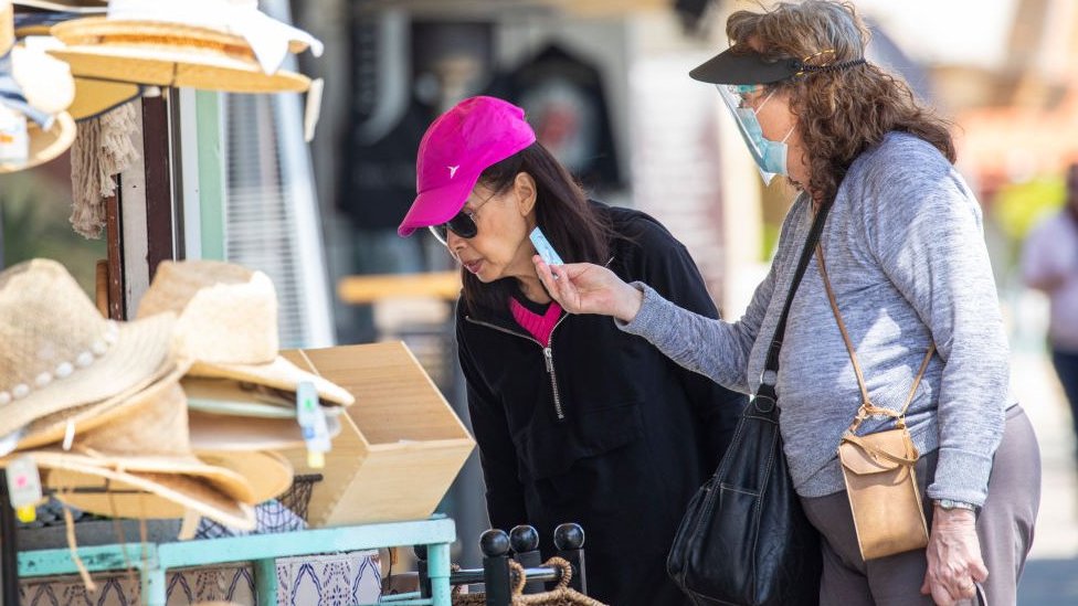 Uma mulher com máscara, e outra sem, olhando produtos na área externa de uma loja