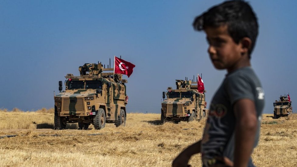 Niño en la frontera de Turquía y Siria con tanques militares en el fondo.