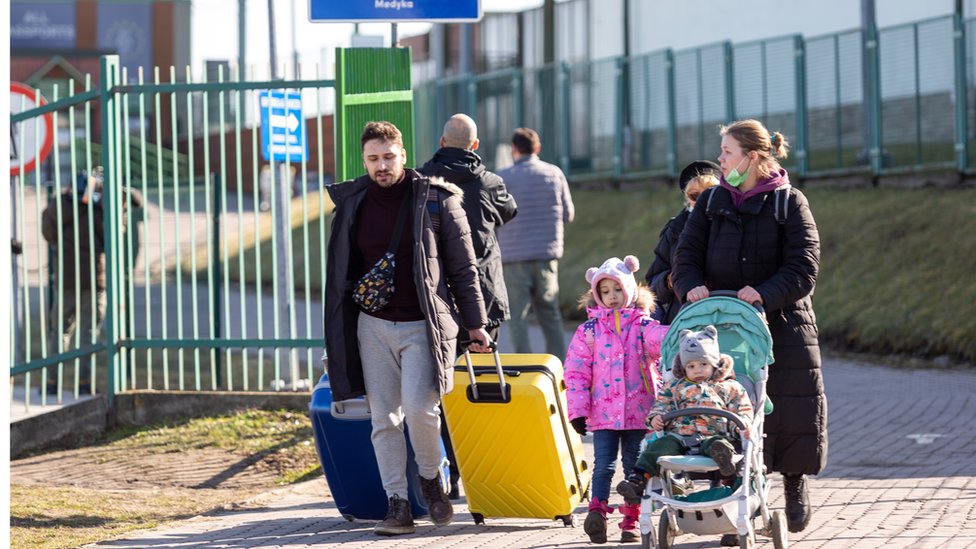 A family with a toddler and a child in a pram, trailing suitcases, cross into Poland