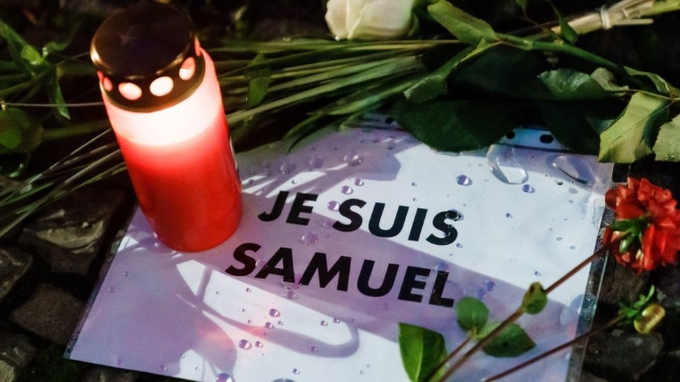 Свеча и цветы оставляют рядом с сообщением на французском языке, которое гласит: «Я Самуэль