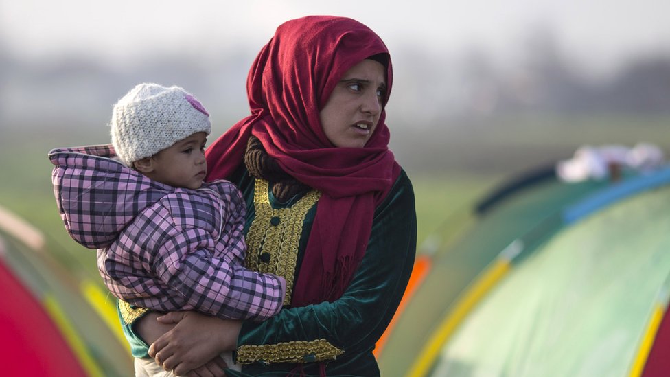 Женщина стоит у своей палатки с ребенком на руках рано утром на границе Греции и Македонии.