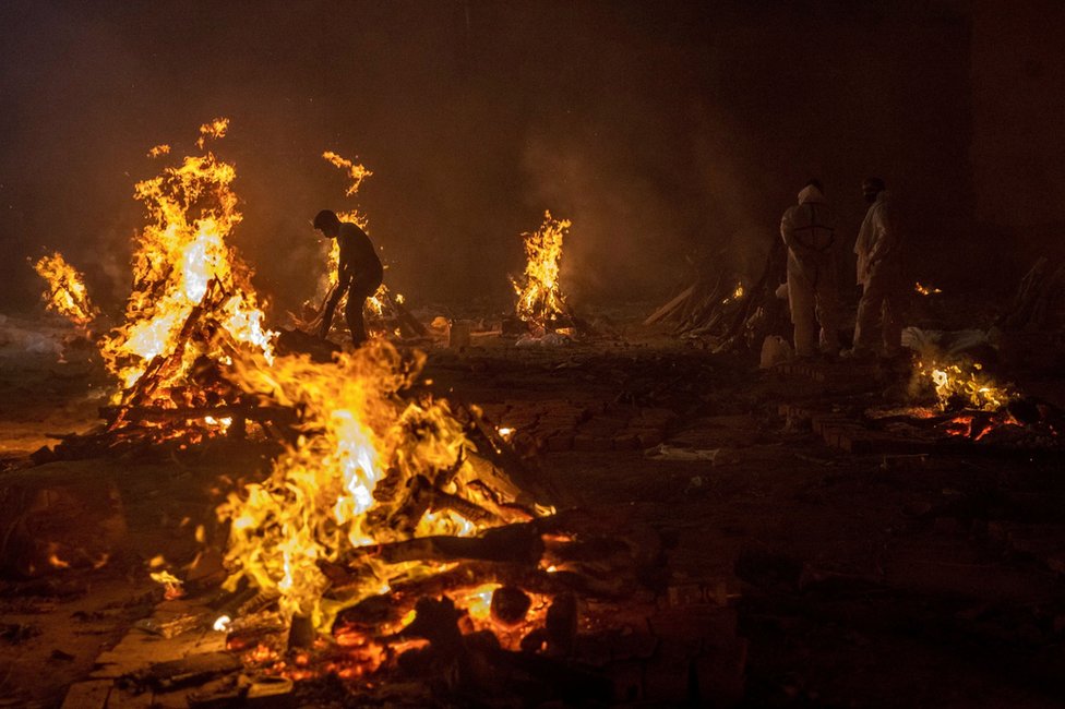 Workers and relatives stand around burning funeral pyres at night