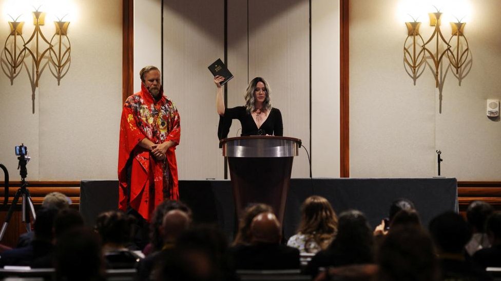 Chalice Blythe, una mujer vestida con un vestido negro, hace gestos con una biblia en la mano durante la apertura de SatanCon.