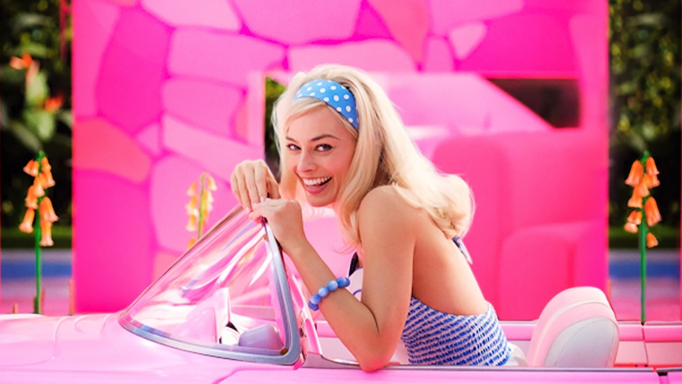 Margot Robbie, uma mulher branca e loira, em um carro rosa