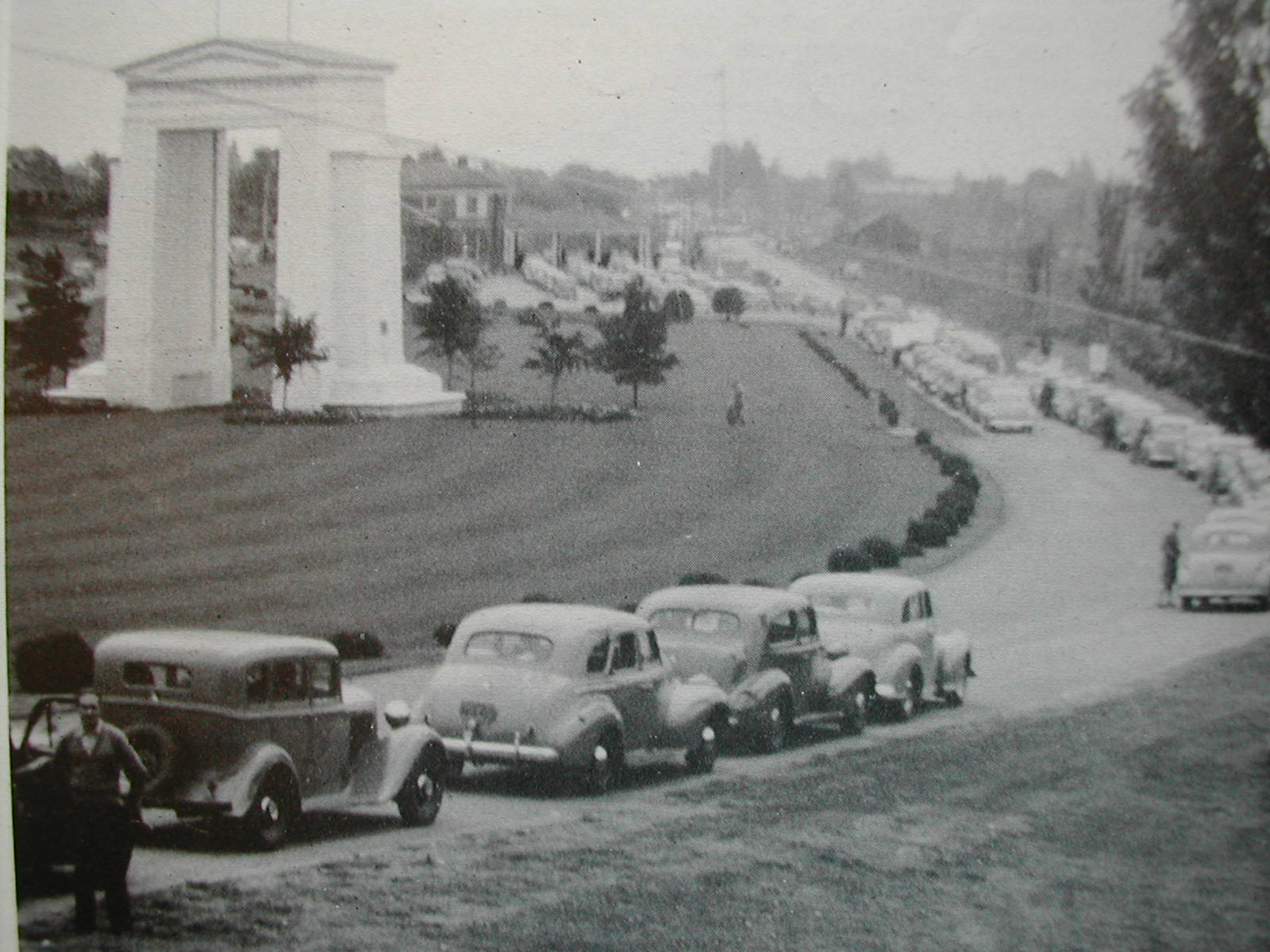 Fotografia em preto e branco mostra diversos veículos em fila ao lado de um arco neoclássico
