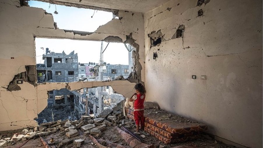 Girl looks out of bombed bedroom wall at a bombed building in Gaza
