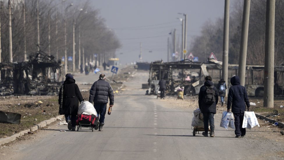 People walking along a street with burnt out vehicles, carrying possessions in bags.
