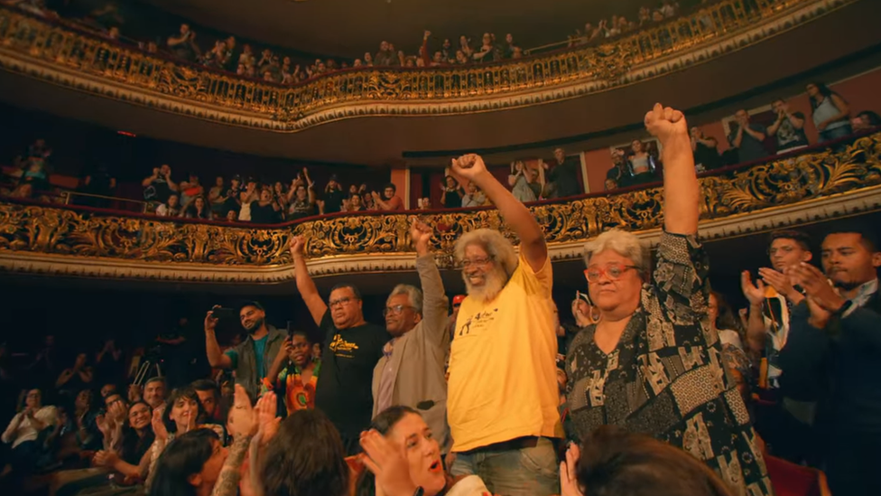 Quatro militantes do MNU, de punhos cerrados, se levantam para receber os aplausos da plateia, durante show do cantor Emicida no Theatro Municipal de São Paulo