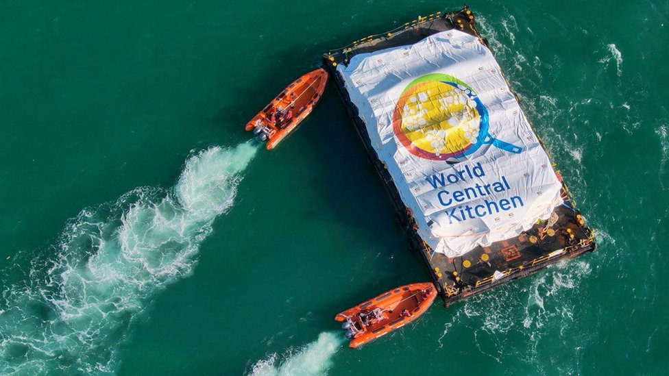 Orange dinghies approach the aid barge. There is a sheet over the aid that reads "World Central Kitchen"