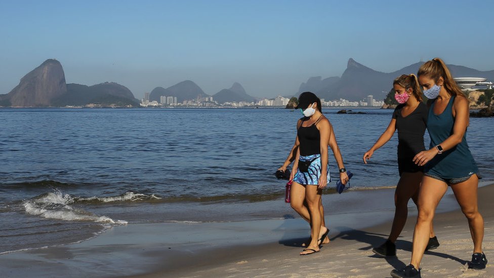 Люди ходят в защитных масках по пляжу Икарай, так как гора Сахарная голова видна вдалеке во время коронавируса (COVID -19) пандемия 21 мая 2020 г. в Нитерой, Бразилия