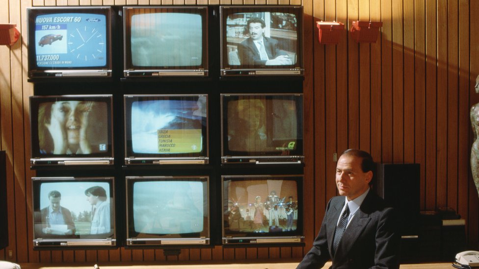 Un Silvio Berlusconi más joven sentadp en una oficina adornada con paneles de madera con nueve televisores muy grandes para la época (1986).