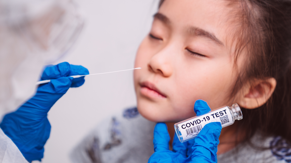 Gloved hands holding a nose swab and a vial labelled 'Covid-19 test', approaching a girl