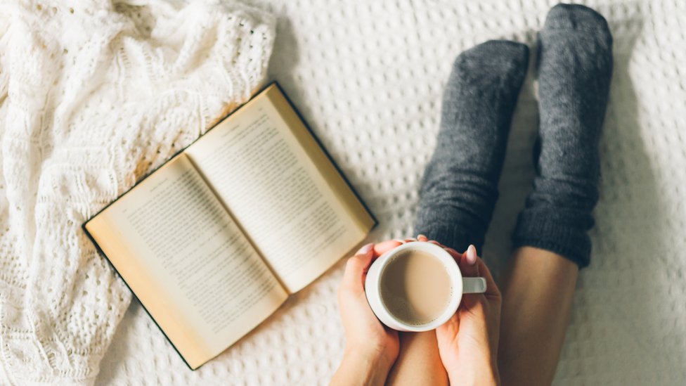 Persona con un libro abierto en la cama y una taza de café.