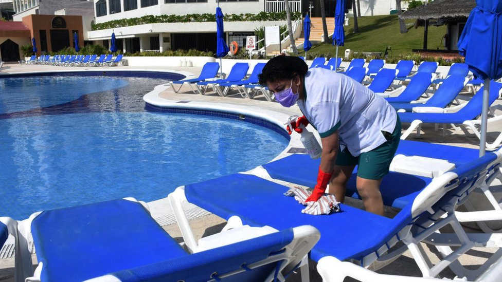 Trabalhadora com máscara limpa espreguiçadeiras no entorno de piscina de resort