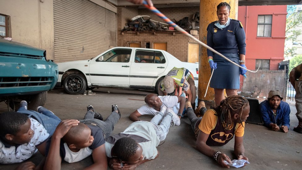 Подозреваемые лежат на полу, пока сотрудник Полицейской службы Южной Африки (SAPS) арестовывает их, потому что они нарушили правила изоляции и были обнаружены с алкоголем в Хиллброу, Йоханнесбург, 27 марта 2020 г.