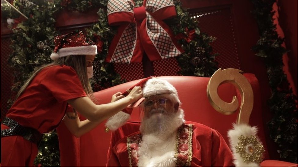 Mulher ajeita homem vestido de Papai Noel usando viseira de plástico