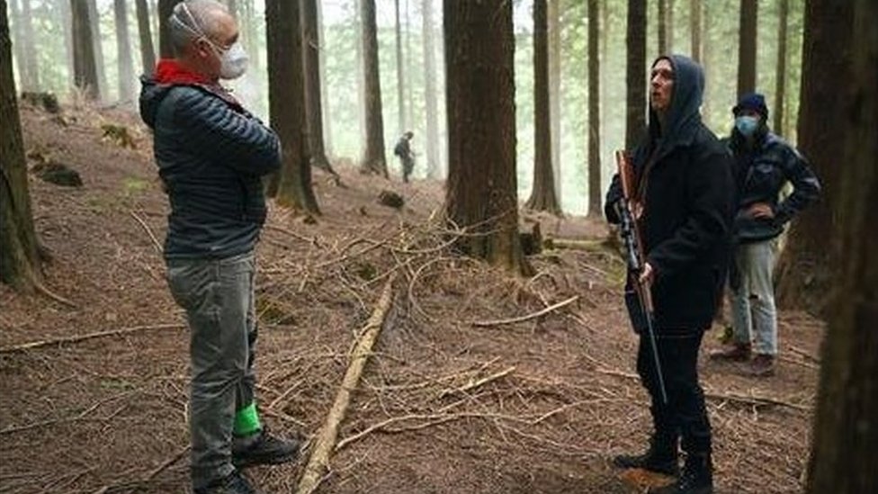 Член экипажа в маске, актер держит фальшивую винтовку в окружении леса