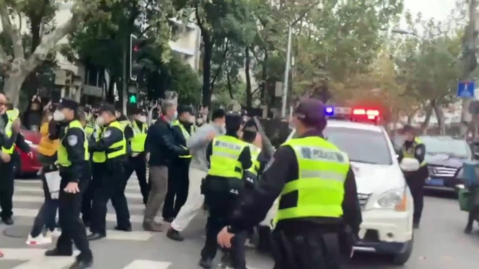 Policiais vestindo jaquetas fluorescentes confrontam manifestantes ao lado de um veículo em Xangai