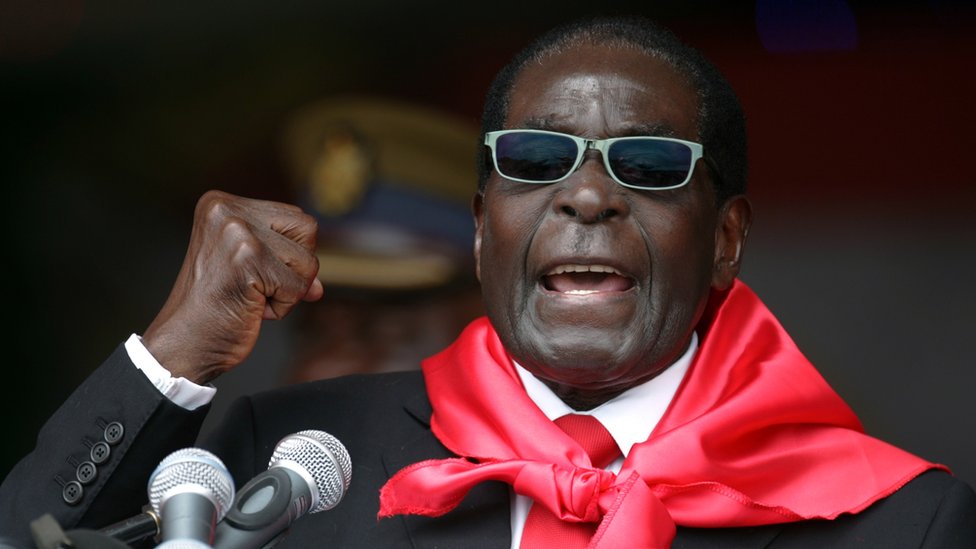Роберт Мугабе, одетый в ярко-красный шарф и солнечные очки, поднимает кулак