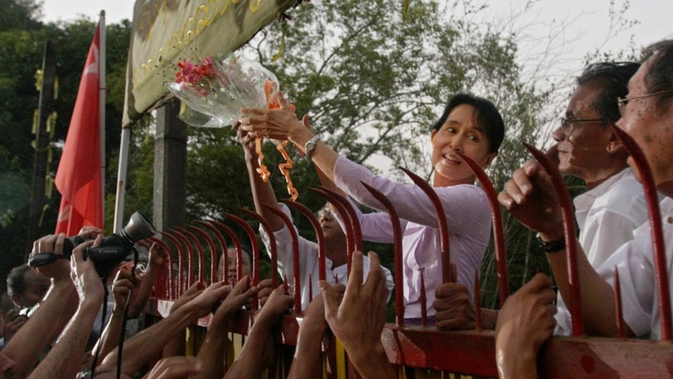 Задержанный лидер оппозиции Мьянмы Аунг Сан Су Чжи держит букет цветов, когда она появляется у ворот своего дома после своего освобождения в Янгоне 13 ноября 2010 года. Лидер демократии Мьянмы Аунг Сан Су Чжи вышла из дома на берегу озера Это была ее тюрьма на протяжении большей части последних двух десятилетий, к радости огромных толп ожидающих ее сторонников. Размахивая руками и улыбаясь, миниатюрная, но неукротимая обладательница Нобелевской премии мира появилась у разрушающегося особняка, в котором она была заперта военной хунтой 15 из последних 21 года.