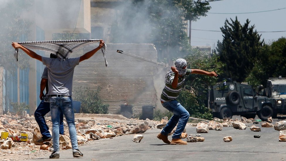 Палестинский протестующий использует рогатку, чтобы бросать камни в израильские силы безопасности во время столкновений после еженедельной демонстрации против экспроприации палестинской земли Израилем 1 июля