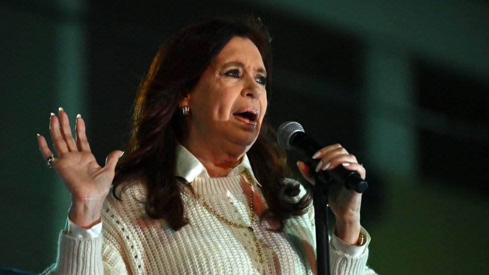 Cristina Fernández, con un micrófono en la mano, la otra palma abierta, mientras da un discurso, abrigada con camisa y jersey blancos.