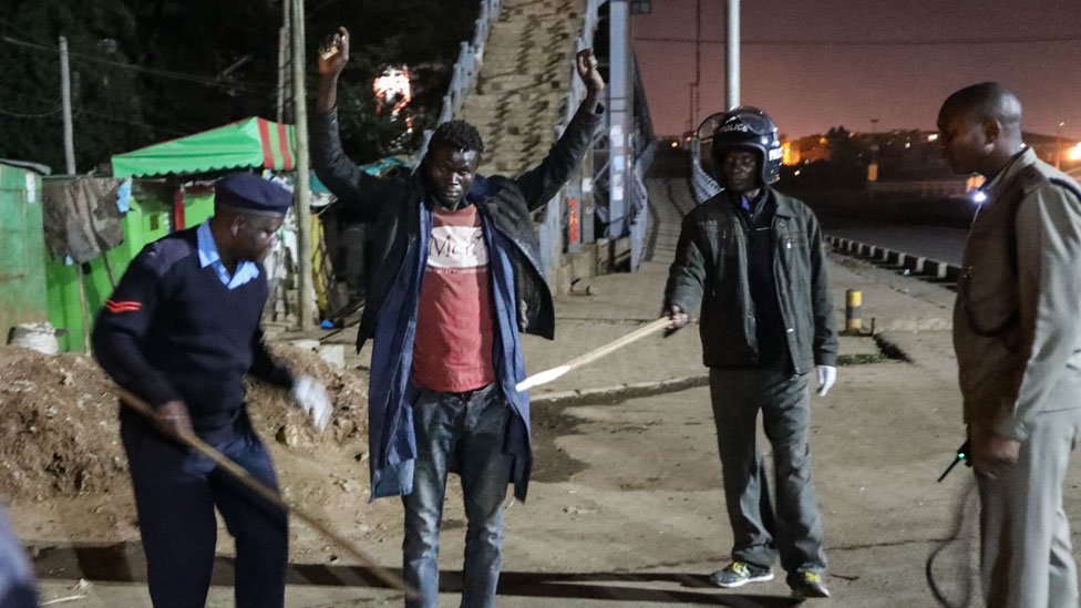 Кенийские полицейские допрашивают и проводят личный досмотр мужчины (С), которого они нашли гуляющим ночью во время патрулирования